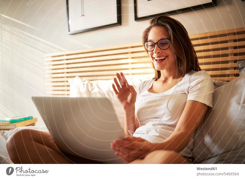 Portrait of laughing young woman sitting on bed with laptop chatting Seated using use Laughter beds females women portrait portraits Laptop Computers laptops