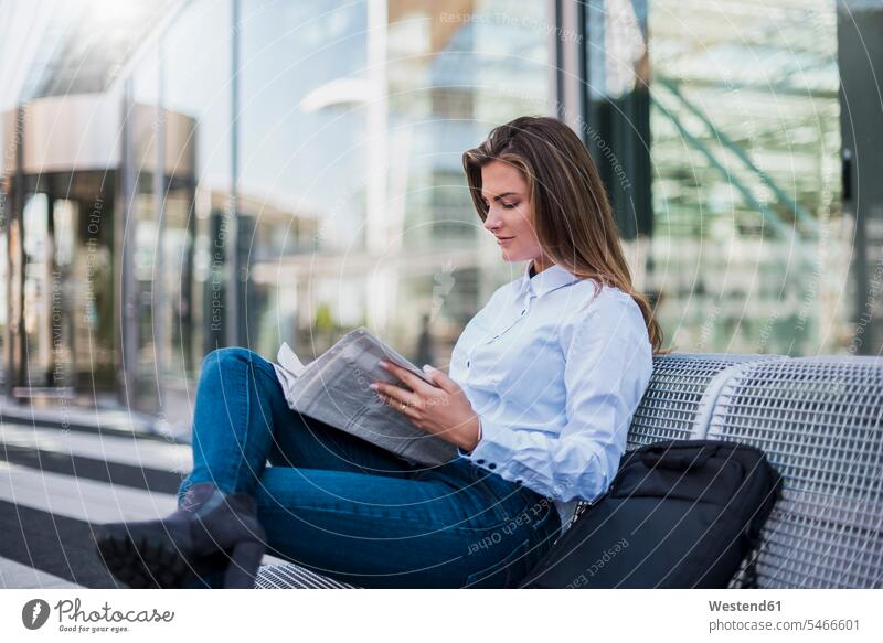 Portrait of young businesswoman sitting on bench reading newspaper businesswomen business woman business women newspapers benches females Seated business people