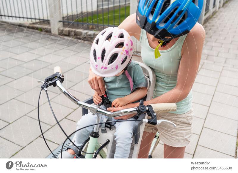 Mother and daughter riding bicycle, daughter wearing helmet sitting in children's seat, fastening seat belt helmets Protective Headwear Child's Seat child seat