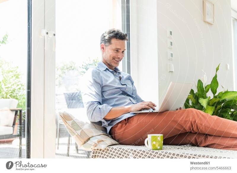Mature man sitting on couch, using laptop windows computers Laptop Computer Laptop Computers laptops notebook smile Seated drink delight enjoyment Pleasant
