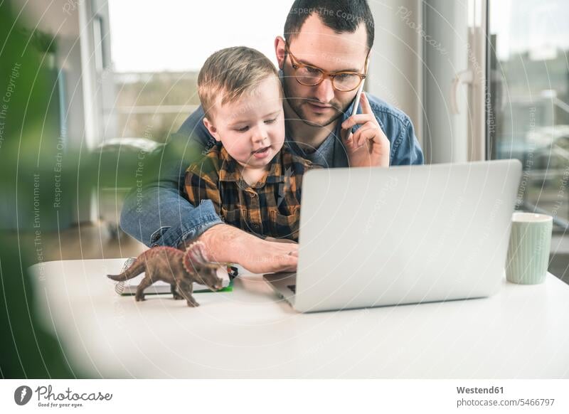 Father working at table in home office with son sitting on his lap Seated sons manchild manchildren working from home home business At Work father pa fathers