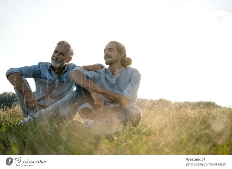 Mature father with adult son sitting on a meadow in the countryside shirts relaxing smile Seated content Contented Emotion pleased country side free time