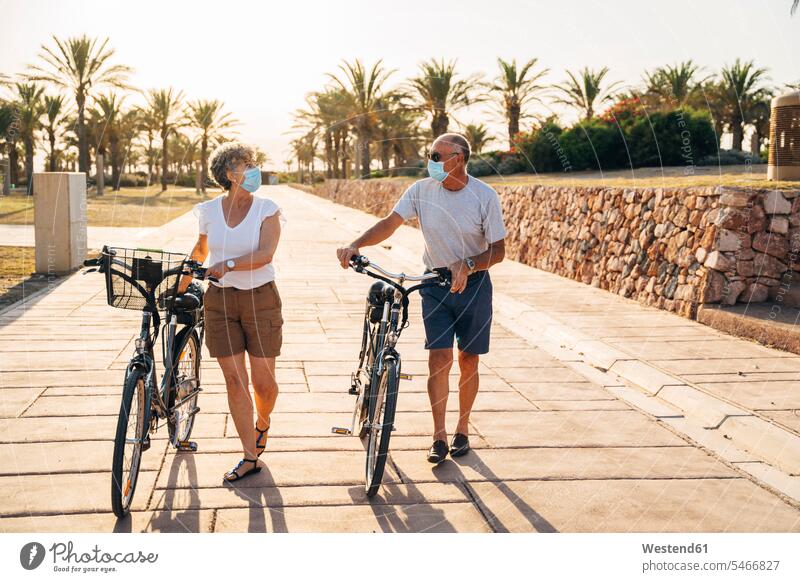 Senior man and woman with face mask talking while walking with cycle at park color image colour image outdoors location shots outdoor shot outdoor shots sunset