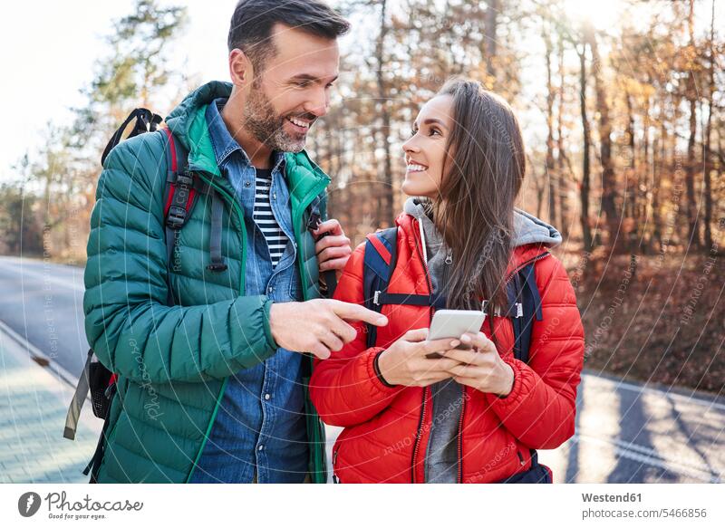 Happy couple checking smartphone on a road in the woods during backpacking trip twosomes partnership couples excursion Getaway Trip Tours Trips rucksacks