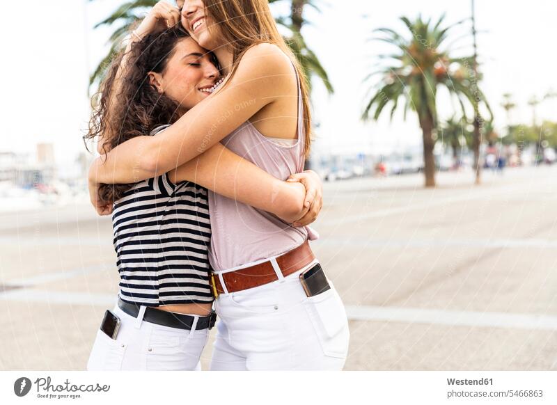 Two happy female friends embracing and hugging on promenade with palms Palm Palm Trees Palms promenades cuddling happiness embrace Embracement mate friendship
