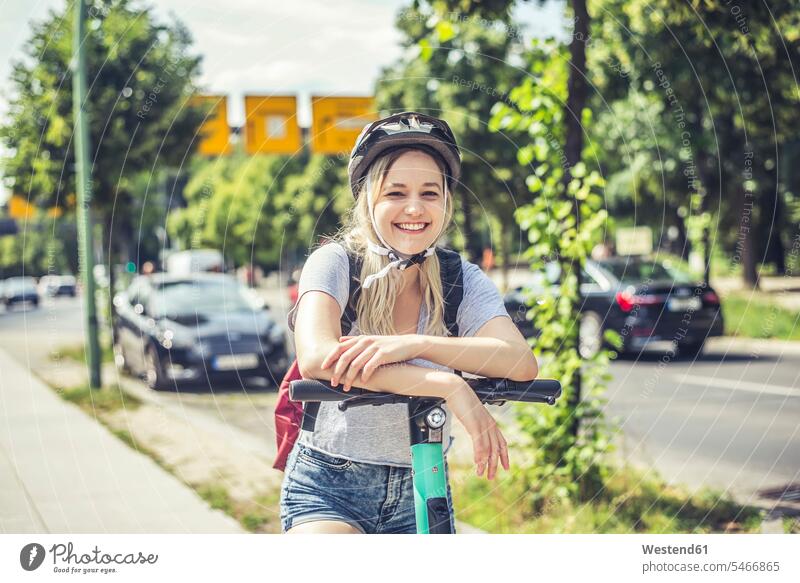 Portrait of happy young woman wearing cycling helmet leaning on handlebar of E-Scooter back-pack back-packs backpacks rucksack rucksacks motor vehicles