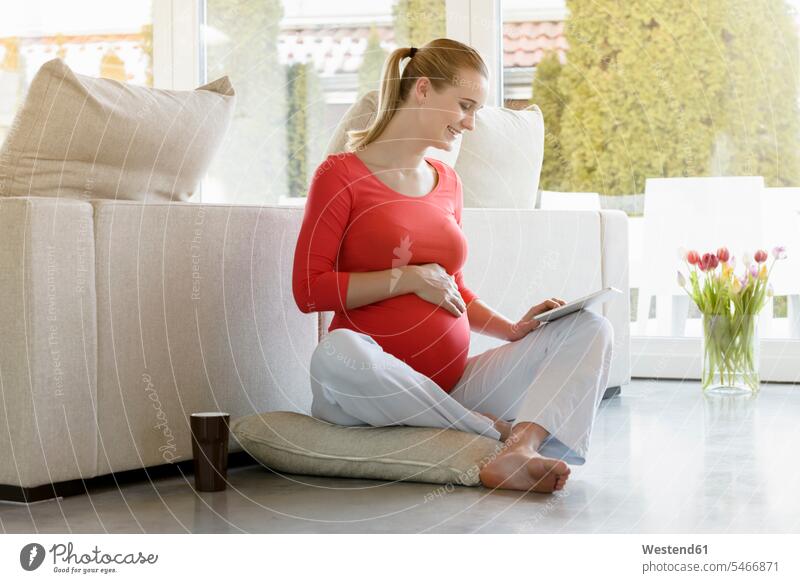 Smiling pregnant woman sitting on floor at home using tablet Pregnant Woman floors smiling smile females women Seated digitizer Tablet Computer Tablet PC