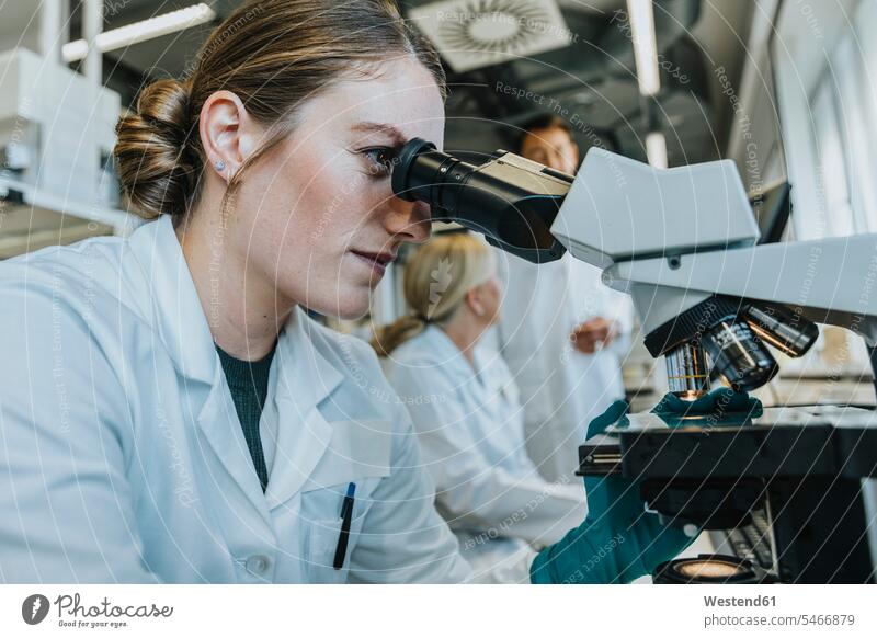 Assistant analyzing human brain microscope slide while sitting with scientist in background at laboratory Incidental people background people