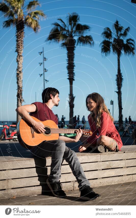 Spain, Barcelona, smiling couple with a guitar sitting on a bench at seaside promenade benches guitars smile promenades twosomes partnership couples Seated