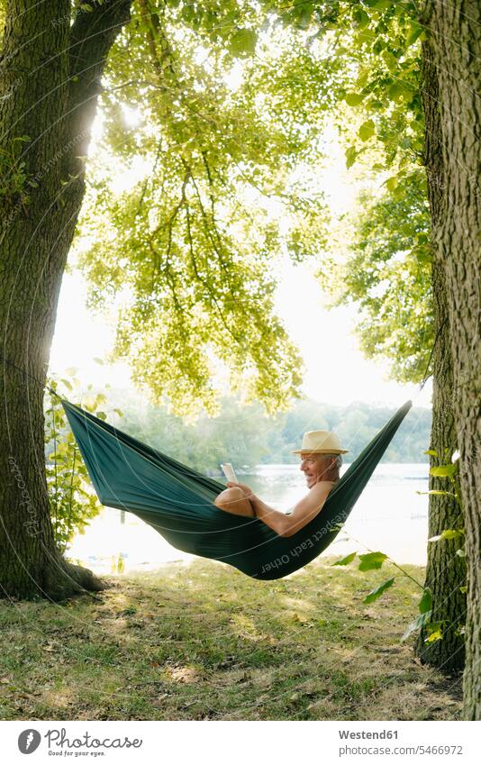 Senior man wearing straw hat relaxing in hammock at lakeshore reading book books Lakeshore Lake Shore lakeside senior men senior man elder man elder men