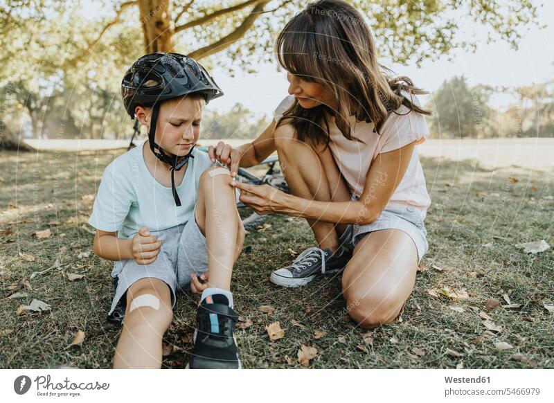 Mother putting bandage on son's knee while sitting in public park during sunny day color image colour image outdoors location shots outdoor shot outdoor shots