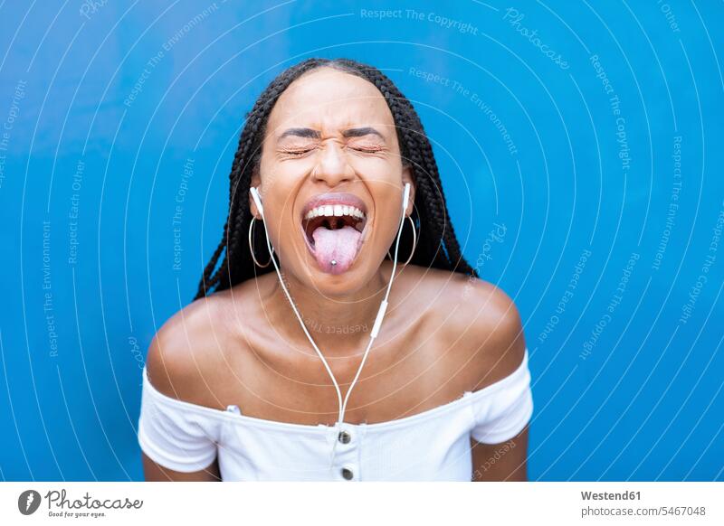 Woman listening music while sticking tongue out against blue wall color image colour image outdoors location shots outdoor shot outdoor shots day daylight shot