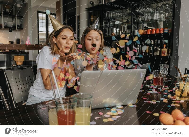 Cheerful friends blowing confetti while video conferencing over laptop while sitting at home color image colour image Germany indoors indoor shot indoor shots