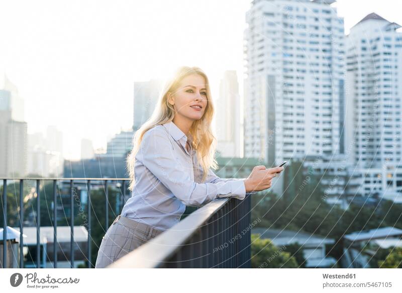 Blonde smiling business woman leaning onto handrail holding smartphone on city rooftop businesswoman businesswomen business women rested on house top Roof Top