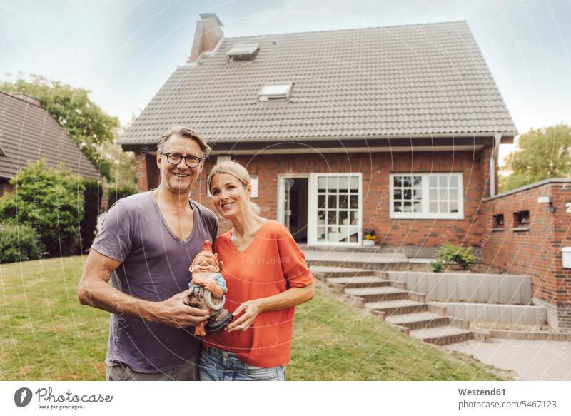Portrait of smiling mature couple standing in front of their home holding garden gnome garden gnomes house houses portrait portraits gardens domestic garden