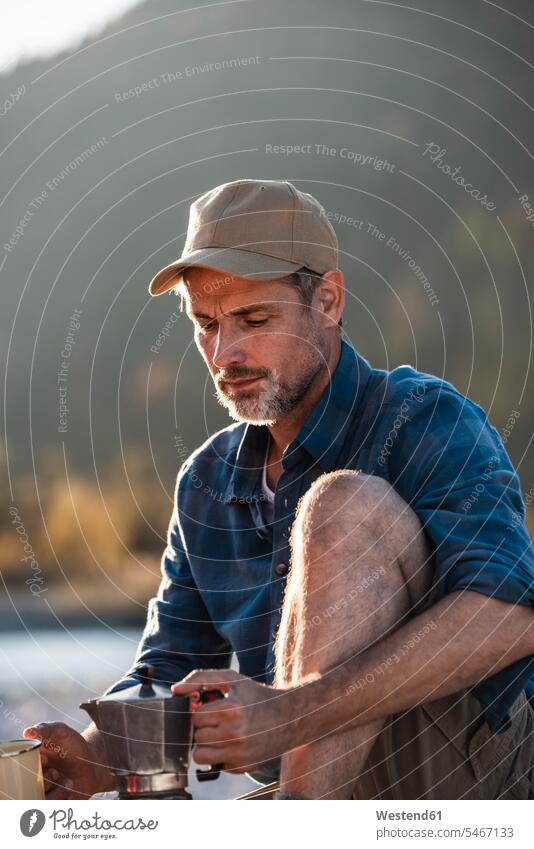 Mature man camping at riverside, using espresso machine baseball cap visor cap peaked cap visored cap Adventure adventurous Adventures nature experience men