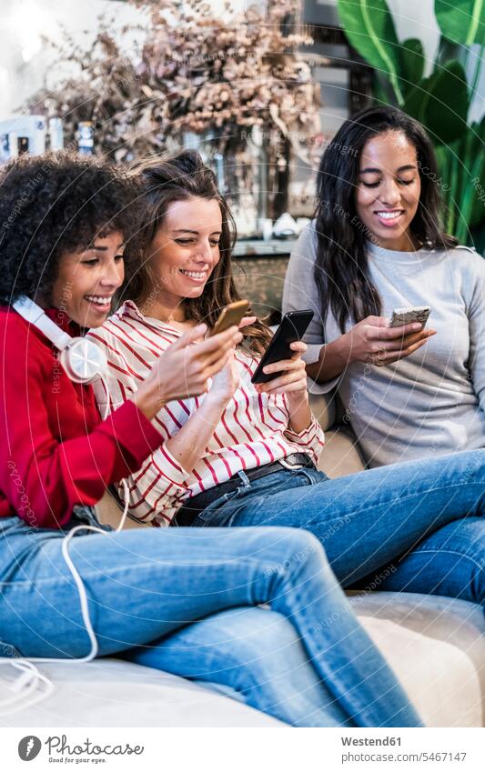 Three smiling women sitting on couch using cell phones mobile phone mobiles mobile phones Cellphone happiness happy woman females Seated female friends settee