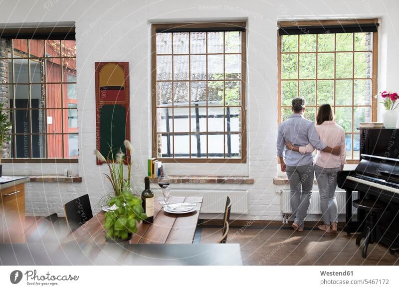 Mature couple standing in their comfortable loft, looking out of window, rear view Germany Home Interior Home Interiors domestic space Looking Through Window