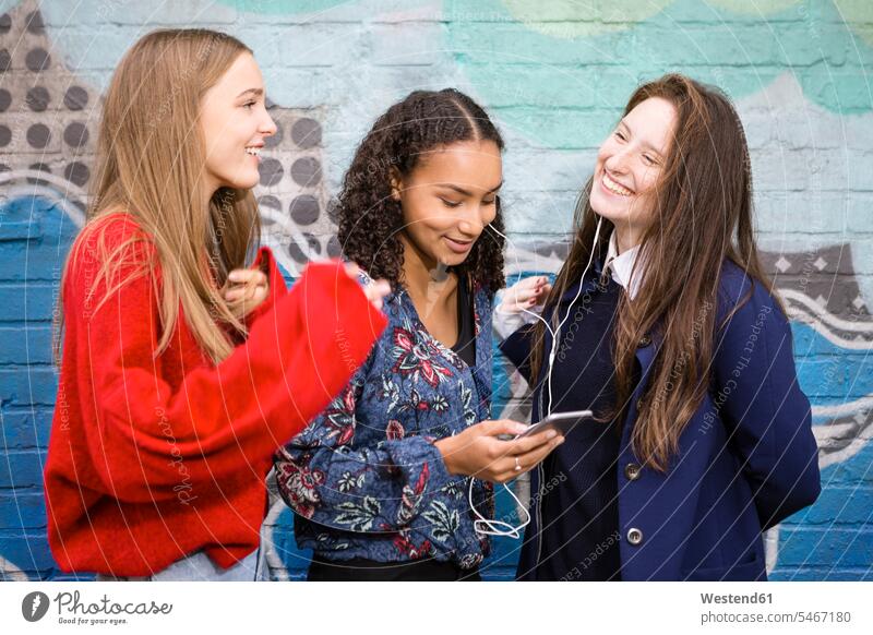 Three friends standing in front of graffiti wall hearing music with smartphone and earphones Graffiti Graffitis female friends ear phone ear phones Smartphone