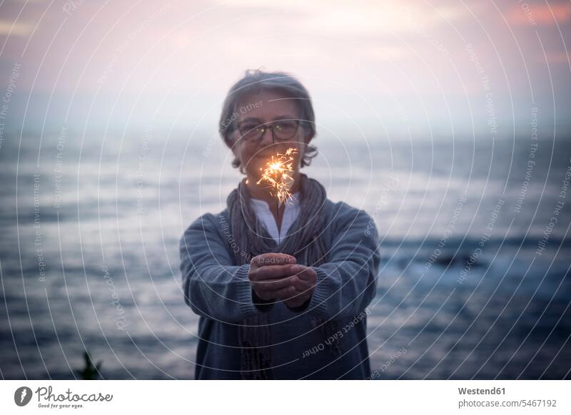 Happy senior woman standing in front of the sea by sunset holding sparkler Spain Moody Sky Romantic Sky sparklers sparks confidence confident relaxation relaxed