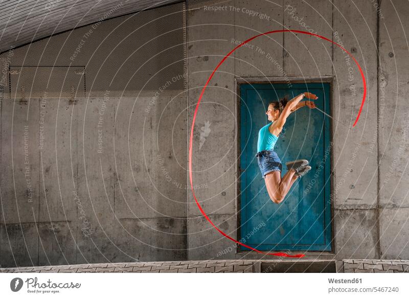 Young woman holding ribbon jumping while doing aerobics against concrete wall color image colour image Germany outdoors location shots outdoor shot