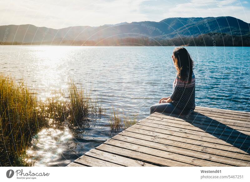 Woman sitting on a wooden platform at a lake at sunset lakes recovering Seated woman females women jetty jetties water waters body of water recreation relaxing