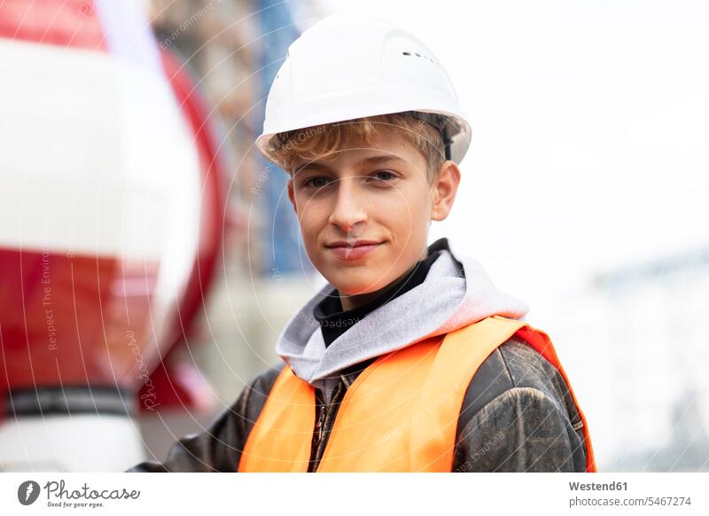 Confident male teenage trainee wearing hardhat and reflective clothing at construction site color image colour image Germany outdoors location shots