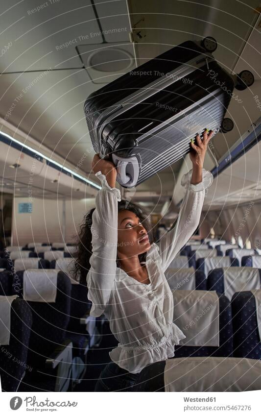 Young woman positioning luggage inside storage compartment in airplane day daylight shot daylight shots day shots daytime casual clothing casual wear