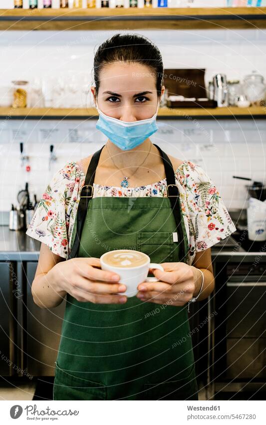 Young woman wearing face mask and apron with coffee cup standing at coffee shop color image colour image indoors indoor shot indoor shots interior interior view