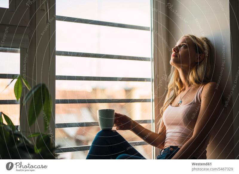 Blond young woman holding coffee mug sitting in windowsill Coffee Mug Coffee Mugs blond blond hair blonde hair Seated looking view seeing viewing window sill