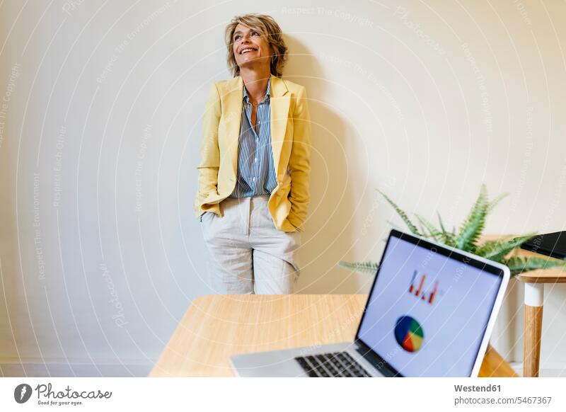 Smiling businesswoman with hands in pockets looking up while standing against wall at office color image colour image indoors indoor shot indoor shots interior