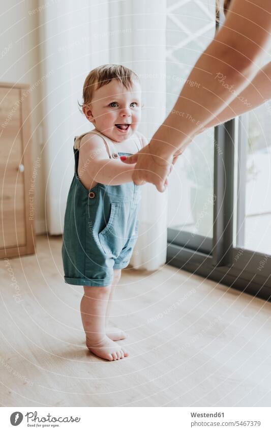 Baby girl holding mother's hands while learning to walk on floor at home color image colour image Spain baby clothing baby clothes Baby Clothings indoors