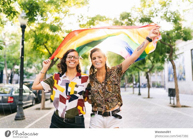 Happy lesbian couple waving rainbow flag while standing on footpath in city color image colour image outdoors location shots outdoor shot outdoor shots day
