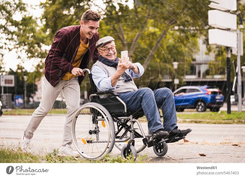 Smiling young man pushing happy senior man with smartphone in wheelchair human human being human beings humans person persons caucasian appearance