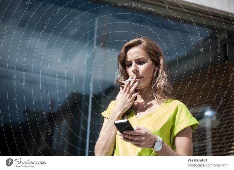 Young female entrepreneur using smart phone while smoking against office building on sunny day color image colour image outdoors location shots outdoor shot