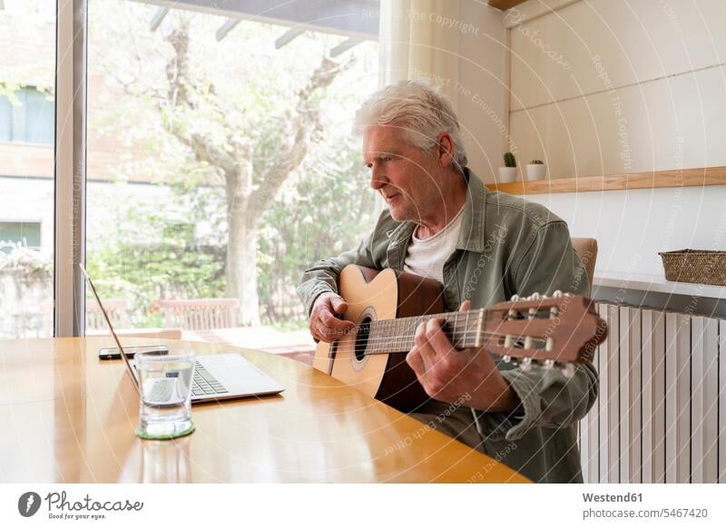Retired elderly man learning to play guitar through online tutorials on laptop at home color image colour image indoors indoor shot indoor shots interior