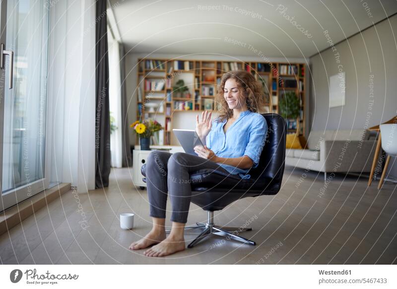 Smiling woman waving hand to video call on digital tablet while sitting on chair at home color image colour image indoors indoor shot indoor shots interior