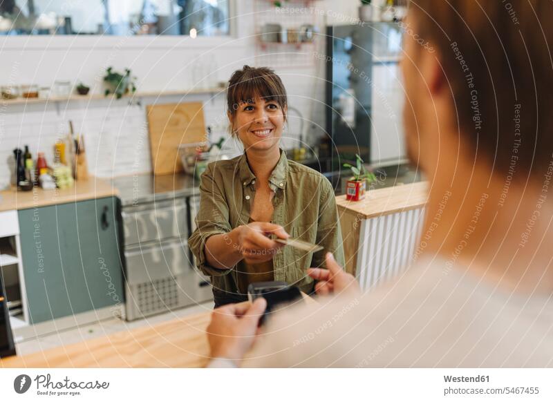 Smiling female owner giving back credit card to male customer at checkout in coffee shop color image colour image indoors indoor shot indoor shots interior