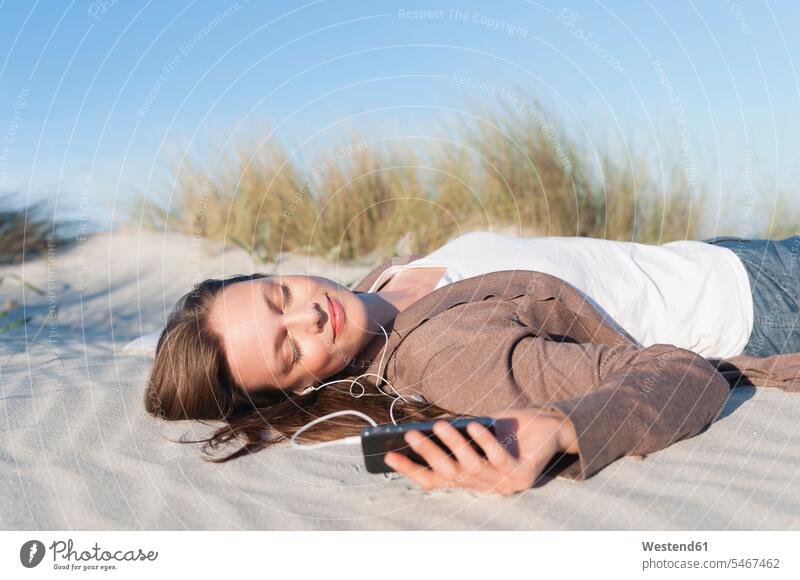 Portrait of woman lying on beach dune listening music with earphones and smartphone, Sardinia, Italy telephone telephones cell phone cell phones Cellphone