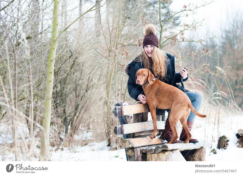 Cheerful young woman sitting on bench with dog against bare trees in forest during winter leisure activity leisure activities free time leisure time one animal