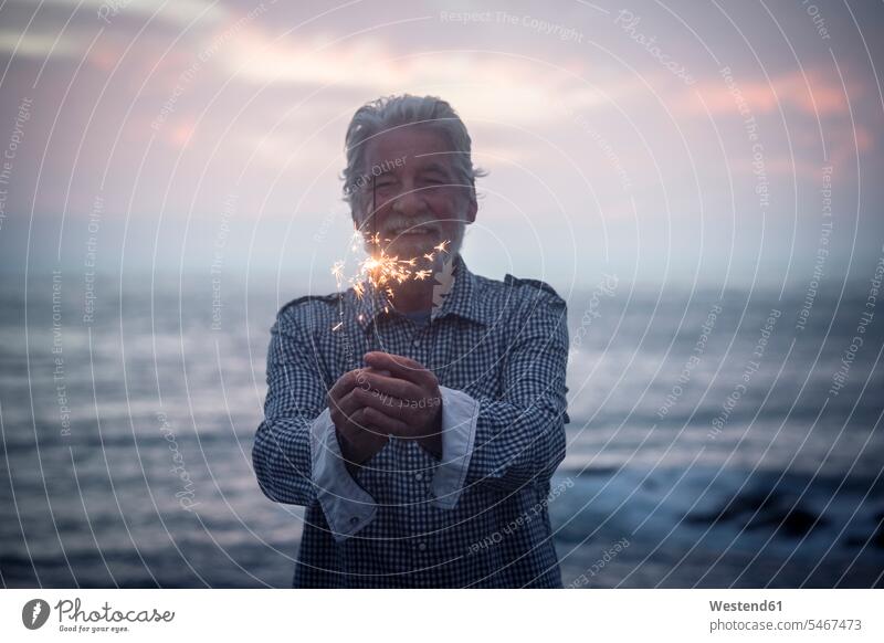 Happy senior man standing in front of the sea by sunset holding sparkler Spain Vignette Moody Sky Romantic Sky sparklers sparks confidence confident copy space