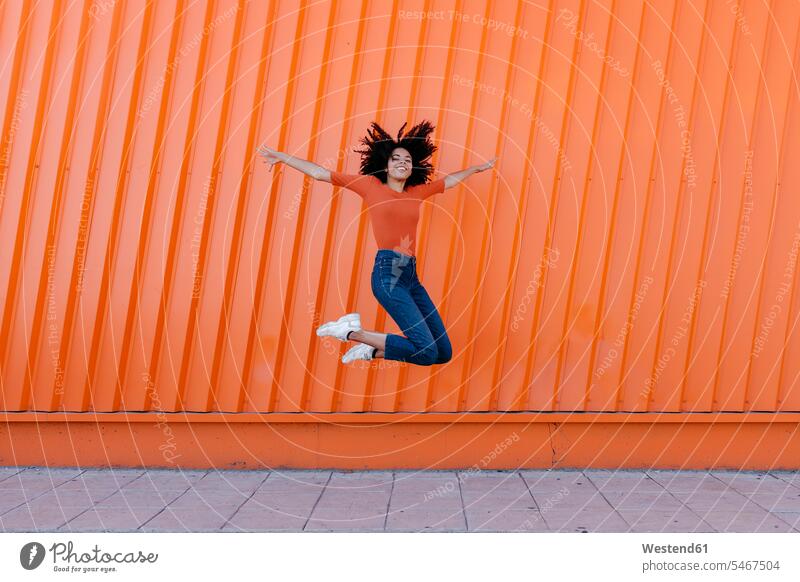 Happy young woman jumping with arms outstretched against orange wall color image colour image outdoors location shots outdoor shot outdoor shots day