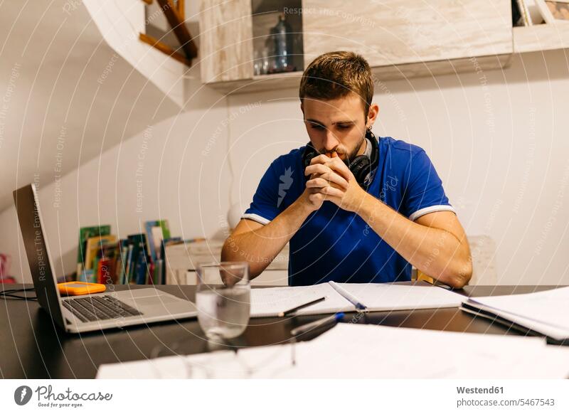 Dedicated young male student sitting with hands clasped while doing homework at table color image colour image indoors indoor shot indoor shots interior