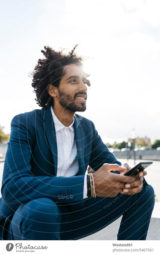 Smiling young businessman sittimg outdoors holding cell phone Businessman Business man Businessmen Business men males sitting Seated smiling smile mobile phone