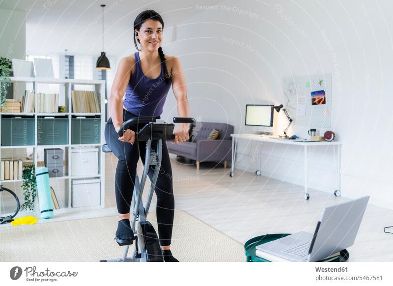 Smiling young woman listening music through bluetooth while sitting on exercise bike at home color image colour image indoors indoor shot indoor shots interior