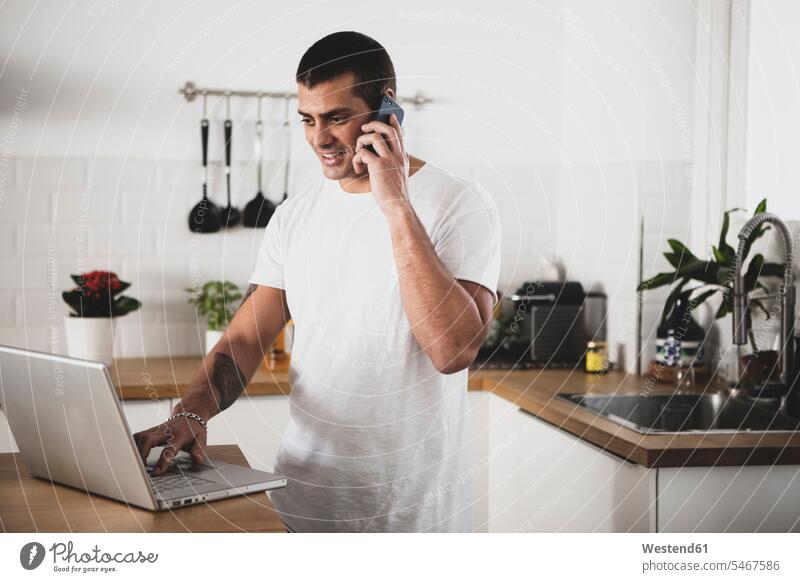 Smiling young man using laptop and cell phone in kitchen at home men males on the phone call telephoning On The Telephone calling smiling smile mobile phone