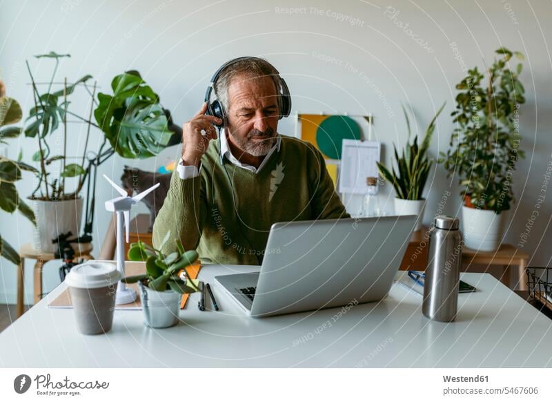 Man wearing headphones using laptop while sitting by table at home color image colour image indoors indoor shot indoor shots interior interior view Interiors