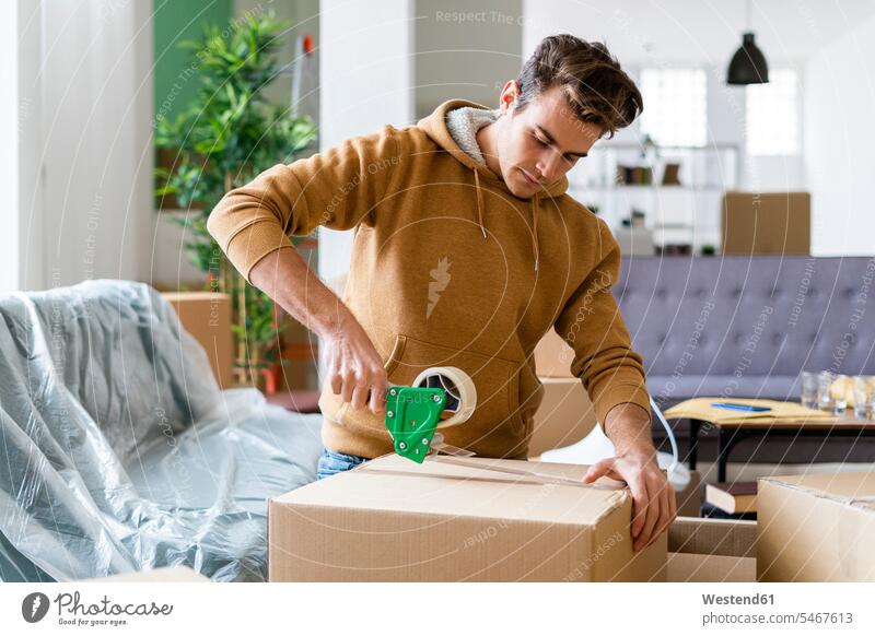 Young man applying adhesive tape on cardboard box while moving in new home color image colour image indoors indoor shot indoor shots interior interior view