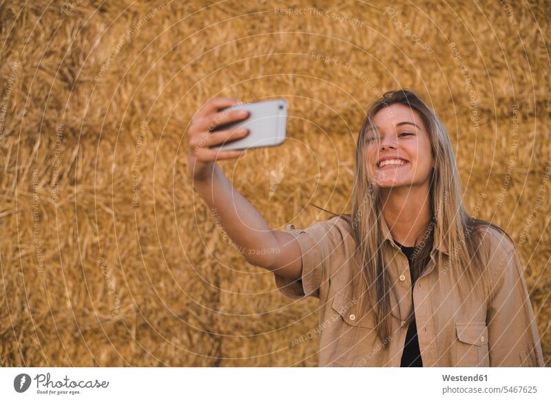Young woman standing in front of hay bales, making smartphone selfie females women Hay beautiful Adults grown-ups grownups adult people persons human being