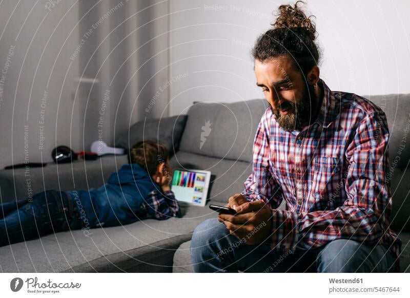 Father sitting on the couch looking at smartphone while his son using digital tablet in the background couches settee settees sofa sofas use seeing view viewing